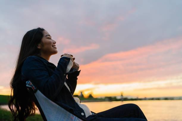 Young woman enjoying hot drink in nature during sunset by lake A young woman is enjoying sitting near a lake and drinking a hot drink in nature during sunset. outdoor lounge stock pictures, royalty-free photos & images